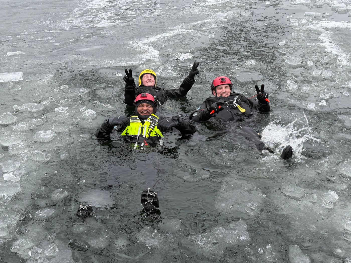 MIPD marine team members in dry suits and helmets floating in icy water during rescue training, smiling and flashing peace signs